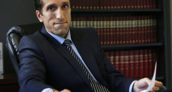 Employment Lawyer Lior Samfiru, Co-founding Partner at Samfiru Tumarkin LLP, sits at a cherry red wooden table with an open binder, leaning back in a leather chair positioned in front of a large legal bookcase, looking confidently at the camera.