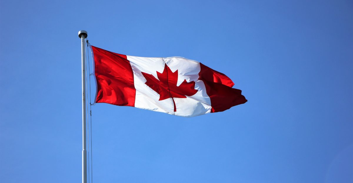 The Canadian flag flies proudly at the top of a flag pole, against a clear, light-blue sky.