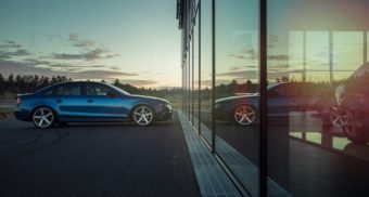 A blue car is parked in front of a reflective glass facade, with a setting sun in the background. Ford Canada employees are entitled to full severance when they lose their job.