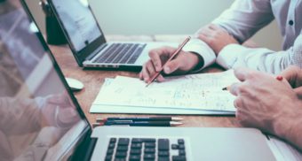 A photo of two people looking at a piece of paper between laptops. (Photo: Scott Graham / Unsplash)