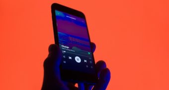 A hand holds up a smartphone that is shown to be playing music, against an orange background. Spotify Canada employees are entitled to severance pay when they lose their job.