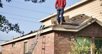 A man in a red sweater is standing on a section of roof that he is in the midst of reshingling. FT Synthetics employees are entitled to severance when they lose their job.