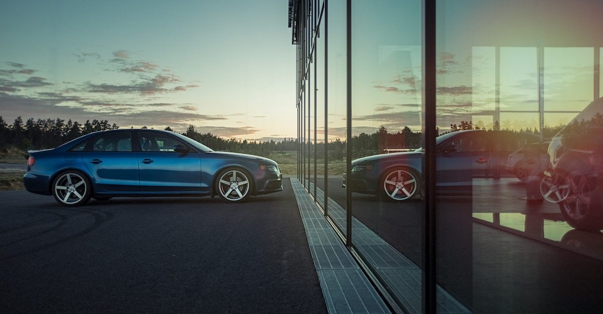 A blue car is parked in front of a reflective glass facade, with a setting sun in the background. Honda Canada employees are entitled to full severance when they lose their job.