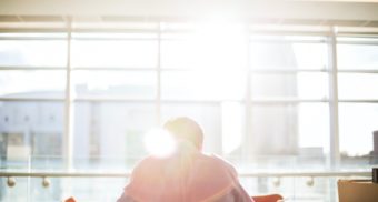 A man bathed in sunlight sits slightly hunched over at an office desk.