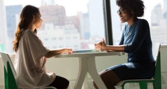 Two women sit at a table for a performance review, with a view of the city skyline through the window next to them.