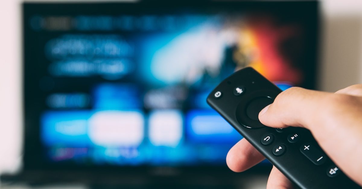 A hand points a remote at an ambiguous streaming service on slightly obscured TV screen. Roku employees are entitled to full severance when they lose their job.