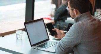 A photo of a person looking at their phone while their laptop loads a webpage. (Photo: Joseph Frank / Unsplash)