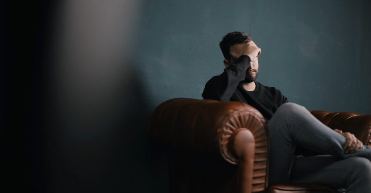 A photo of a person sitting on a brown couch with their hand over their face. (Photo: Nik Shuliahin / Unsplash)