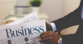 A black man sporting a watch and business suit reads the business section of a newspaper. He recently negotiated an executive employment contract.