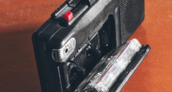 A black, handheld tape recorder sits open on a wooden table with a cassette tap loaded. It is legal to record conversations at work in British Columbia, in some cases.