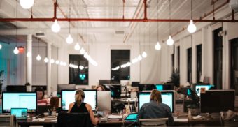 A number of employees are seated at a row of computers in a large workspace, with lightbulbs hanging overhead.