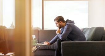 A man in a hoodie sits on a couch while performing sedentary work on a computer.