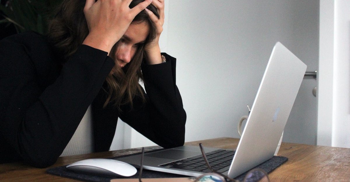 A stressed young woman, an employee working in Ontario, clutches her head as she stares at her laptop screen.