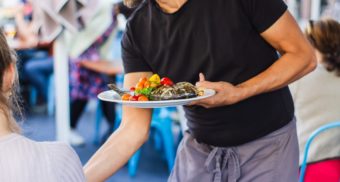 A restaurant server in a black t-shirt delivers a plate of food to a customer's table.