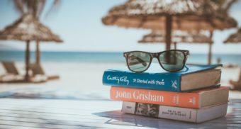 Three books with a pair of sunglasses on top sit on a table with beach and umbrellas in the background.