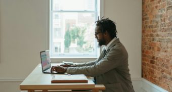 A man sitting at a wooden table in a room in his home, working remotely on his laptop.