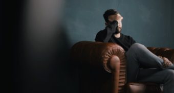 A photo of a person sitting on a brown couch with their hand over their face. (Photo: Nik Shuliahin / Unsplash)