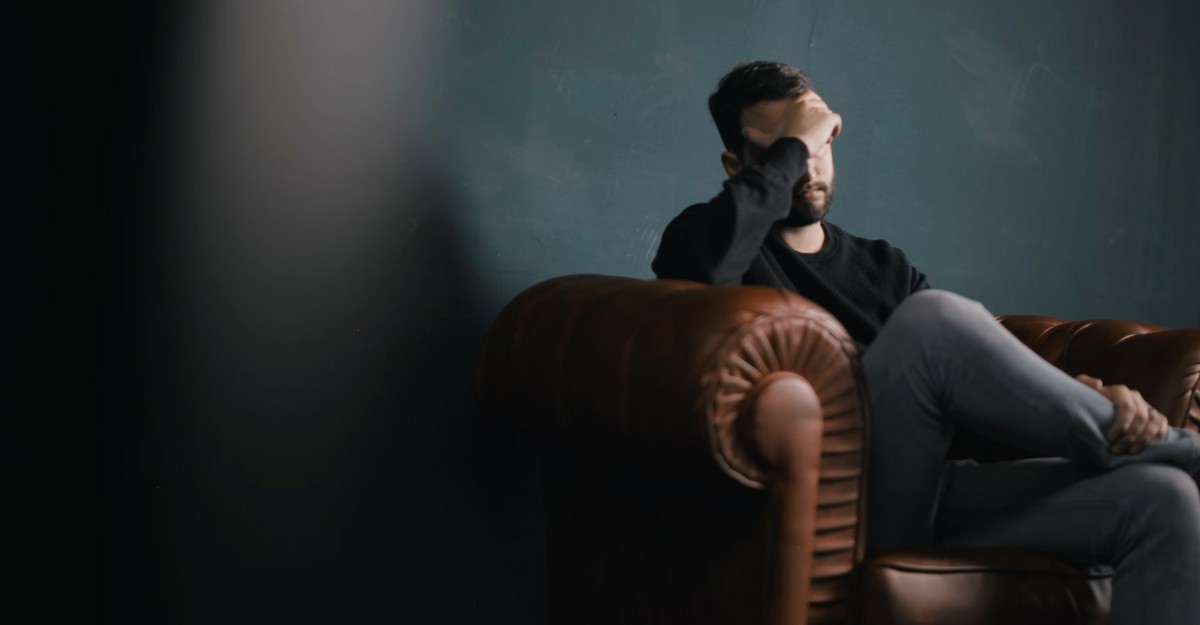 A photo of a person sitting on a brown couch with their hand over their face. (Photo: Nik Shuliahin / Unsplash)