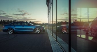 A blue car is parked in front of a reflective glass facade, with a setting sun in the background. WEINS Auto Group employees are entitled to full severance when they lose their job.