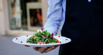 A food server, potentially one provided by Compass Group Canada, holds a plate of food in their hand.