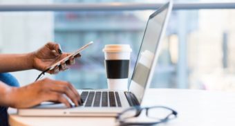 A photo of a person checking their phone while typing on a laptop. (Photo: Wocintechchat / Unsplash)