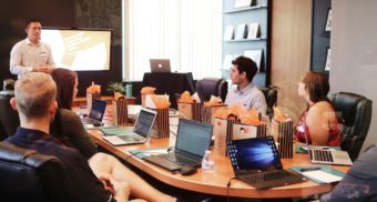 A group of employees are gathered around a large office table, listening to input from the marketing manager.