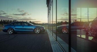 A blue car is parked in front of a reflective glass facade, possibly at a Myers Automotive Group dealership, with a setting sun in the background.