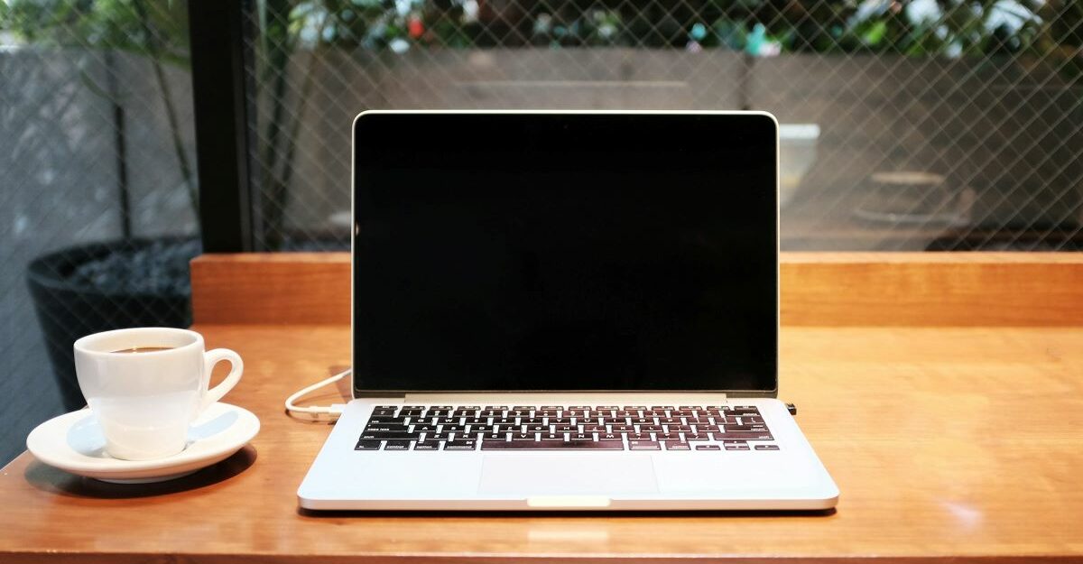 An open laptop, potentially made by Apple, sits on a desk next to a cup of coffee.