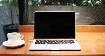 An open laptop, potentially made by Apple, sits on a desk next to a cup of coffee.