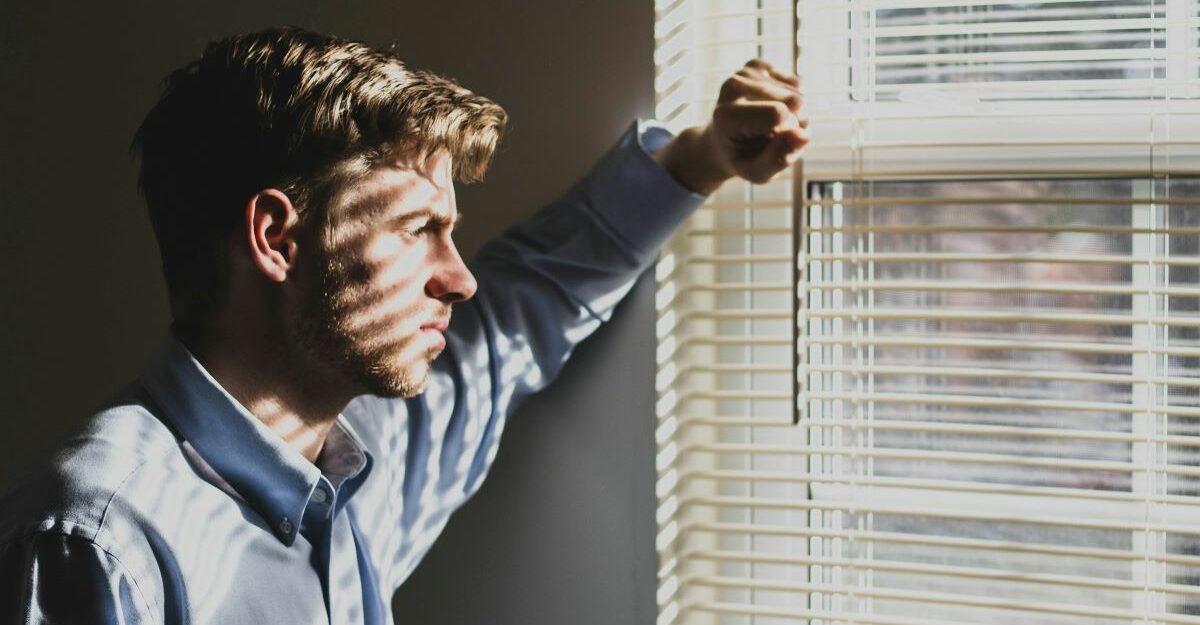 A man with a troubled look on his face stares out through the blinds of a window.