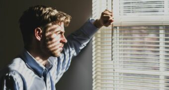 A man with a troubled look on his face stares out through the blinds of a window.