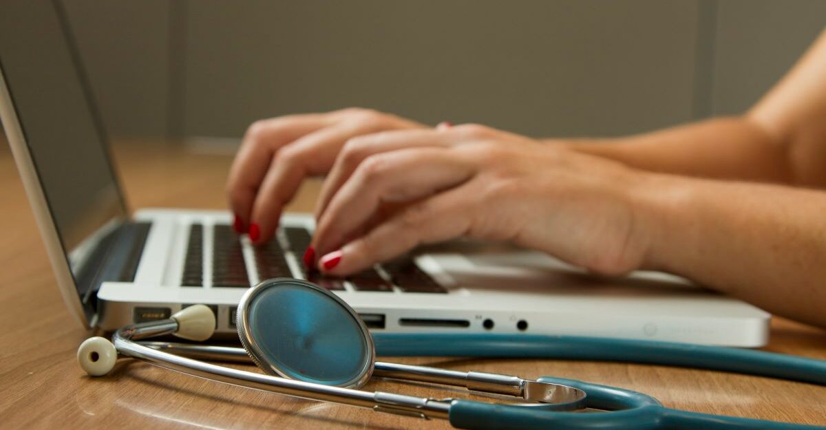 A stethoscope sits on a table next to a person, possibly working for Intuitive Surgical, using a laptop.