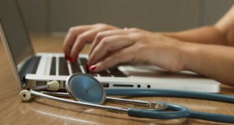 A photo of a person using a laptop with a stethoscope beside them. (Photo: National Cancer Institute / Unsplash)