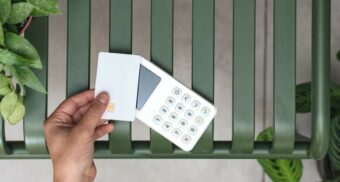 A photo of a person holding a credit card over a payment machine on a bench. (Photo: Mockuuups / Unsplash)