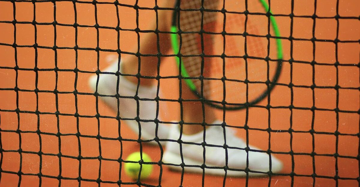 A tennis play stands next to a tennis ball, as seen through the net dividing the court.