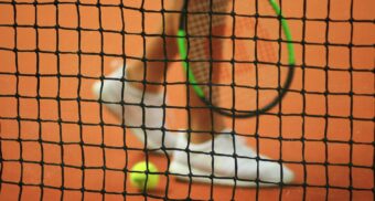 A tennis play stands next to a tennis ball, as seen through the net dividing the court.