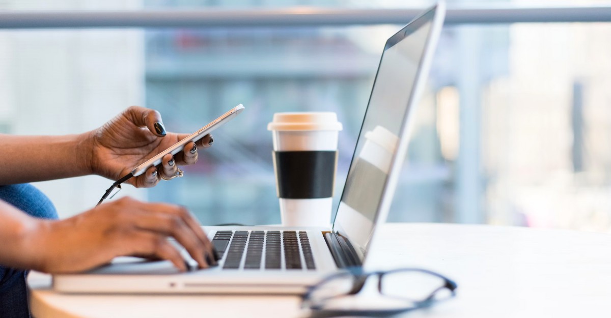 A photo of a person checking their phone while typing on a laptop. (Photo: Wocintechchat / Unsplash)