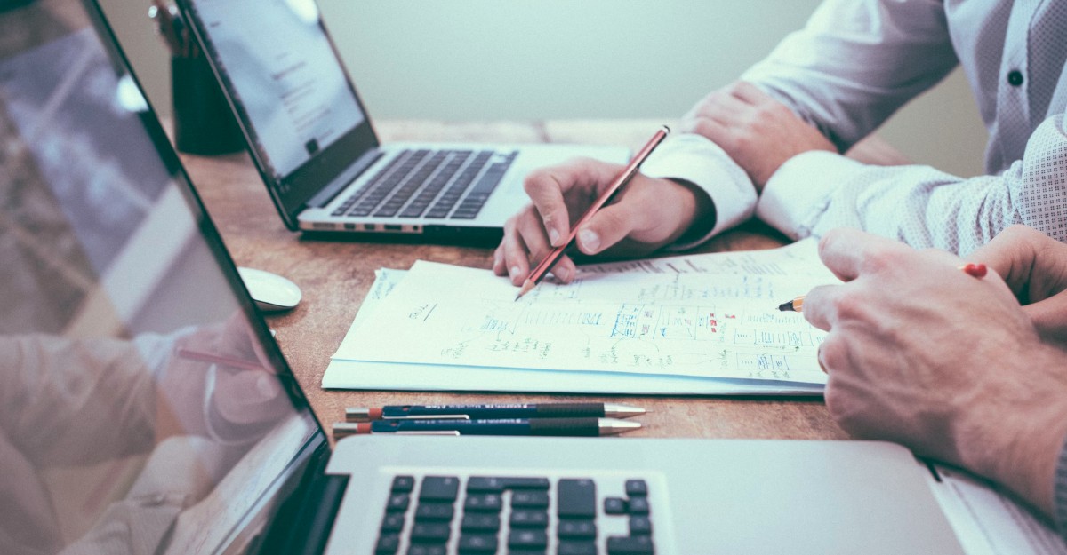 A photo of two people looking at a piece of paper between laptops. (Photo: Scott Graham / Unsplash)