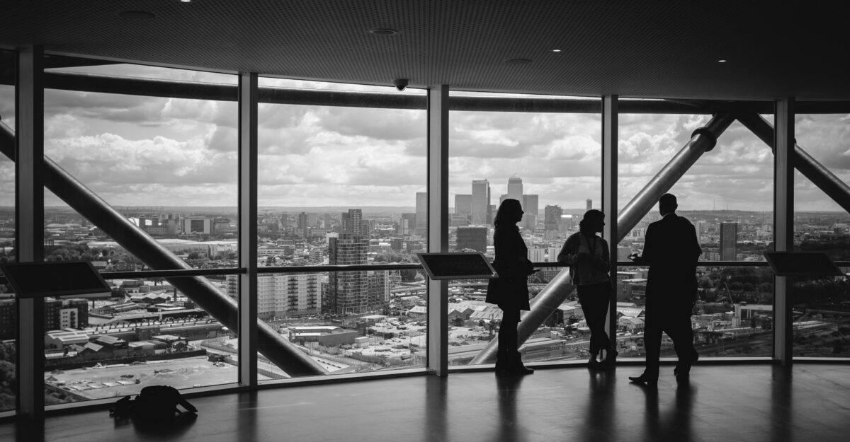 Three people discuss business while standing next to a window with a view of a city.