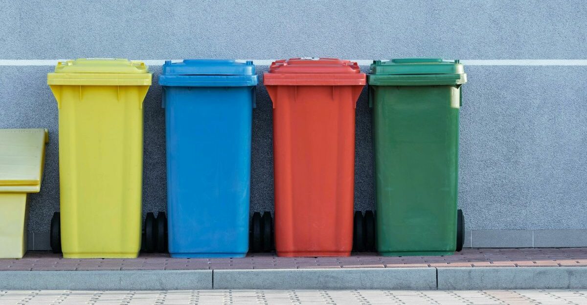 Yellow, blue, red, and green recycling bins are lined up against a wall. (Unsplash)