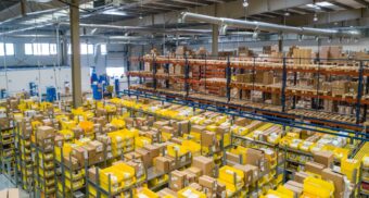 Interior view of a large warehouse with shelves full of cardboard boxes and yellow storage bins, representing an Amazon fulfillment centre.