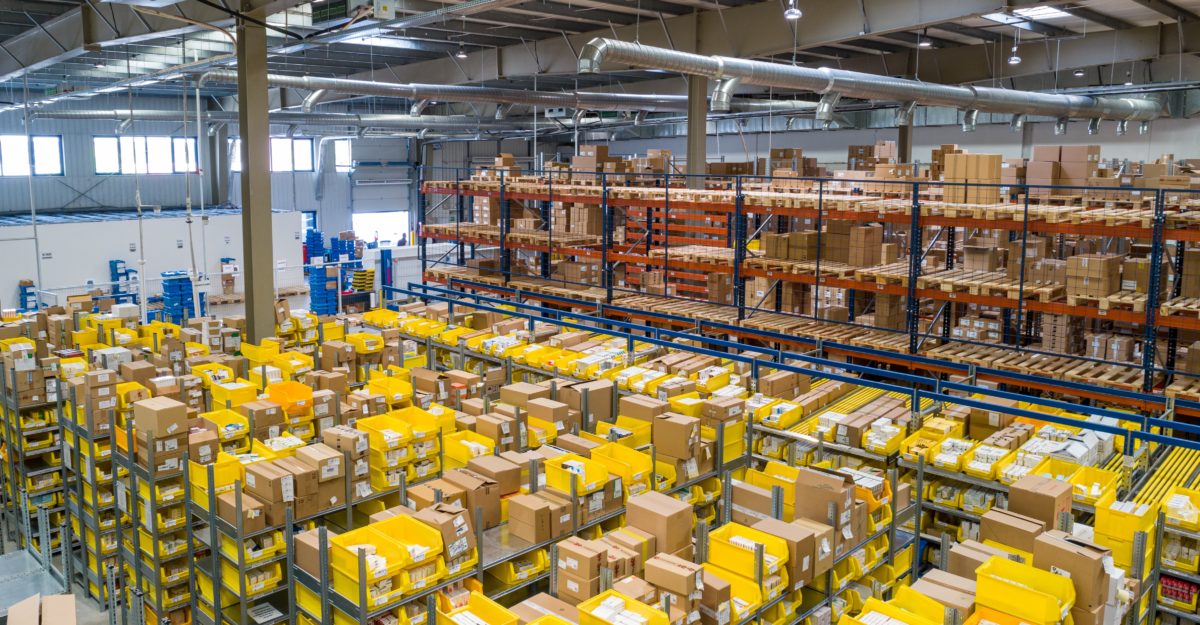 Interior view of a large warehouse with shelves full of cardboard boxes and yellow storage bins, representing an Amazon fulfillment centre.
