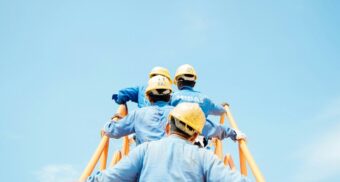 A group of construction workers in blue uniforms and yellow helmets climbing scaffolding under a clear blue sky, symbolizing dependent contractors in Ontario who rely on one main company for work.