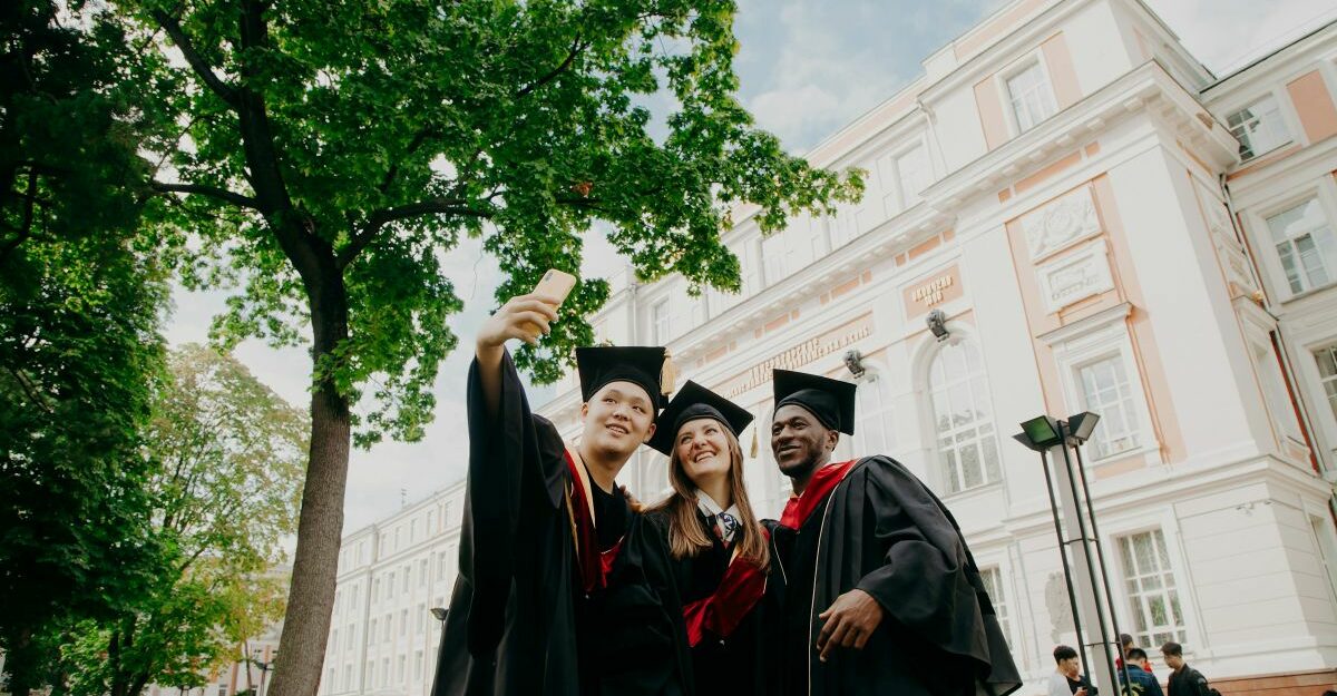 A trio of international students pose for a group selfie following a graduation ceremony at a college or university in Canada.