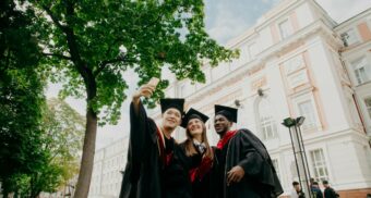 A trio of international students pose for a group selfie following a graduation ceremony at a college or university in Canada.