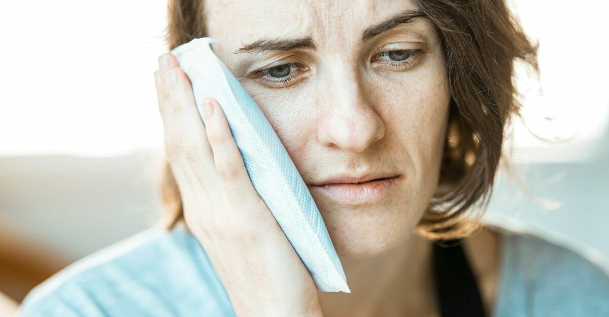 A woman holding an ice pack to her face, appearing to be in pain and discomfort.
