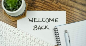 A desk with a keyboard, notebook, pen, and a small plant surrounding a paper sign that says “Welcome Back.”