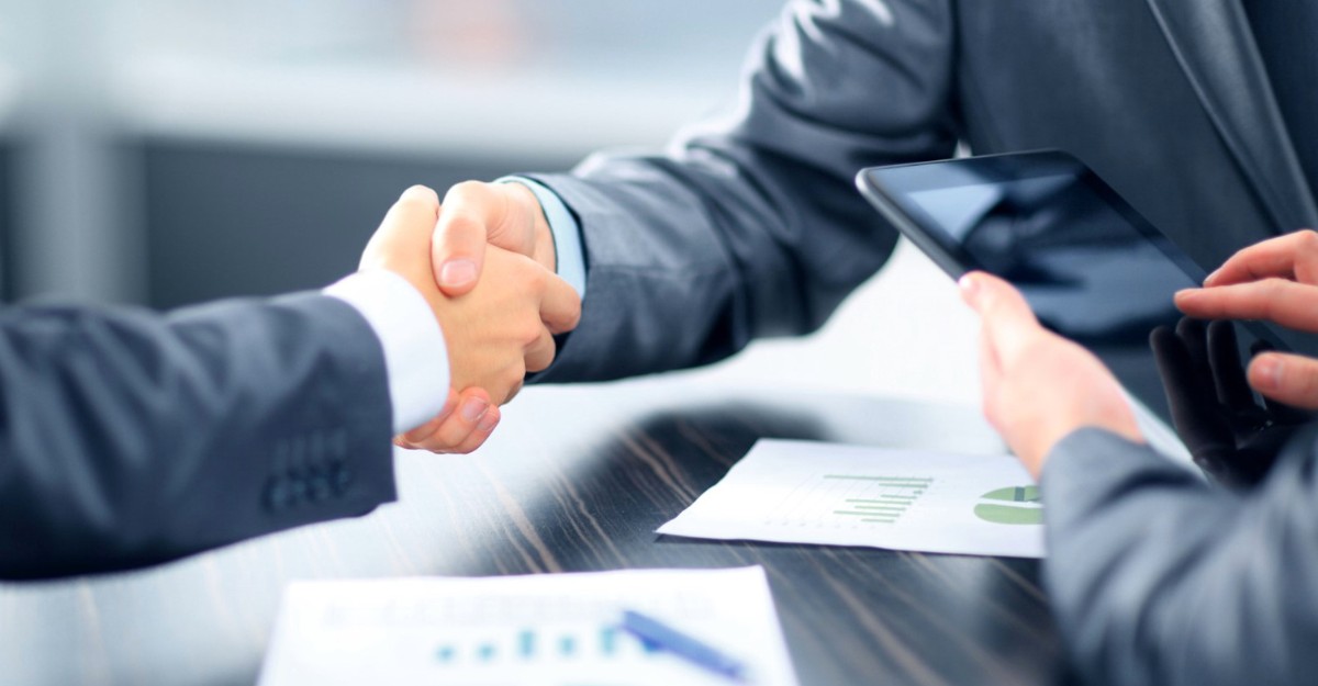 Two people shaking hands while a third person reviews a tablet. (Photo: business-to-you.com)
