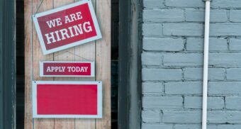 A red sign hangs on the door of a business, revealing that they are hiring a new employee.